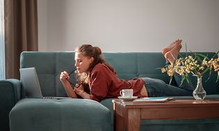 Kvinna ligger på en gråblå soffa med en bärbar dator framför sig och en tekopp bredvid sig på soffbordet