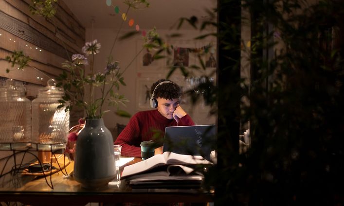 Ung manlig student sitter hemma framför sin dator med många böcker uppslagna framför sig och hörlurar på