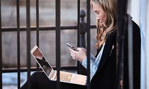 Ung kvinna sitter utomhus med en jacka runt sig med en iphone och uppslagen macbook