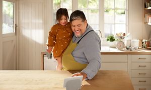 Glad man med förkläde håller en liten flicka medan de tittar på en skärm som står på en köksö med träbänkskiva i ett ljust kök