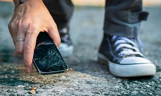 En person plockar upp sin mobiltelefon med krossad skärm från marken.