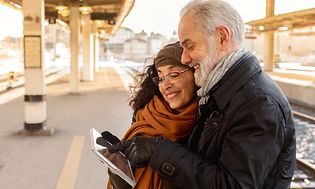 An older couple standing on a train platform looking at a tablet