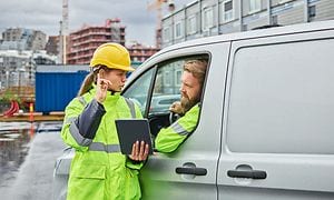 Kvinna i arbetskläder med en surfplatta på en byggarbetsplats pratar med en man i en bil med arbetskläder
