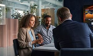 En kvinna och två män sitter vid ett bord i ett mötesrum med glasvägg och statistik på en skärm