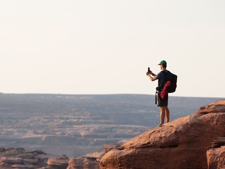 Man with caps and shorts on a cliff taking pictures of the view with his smartphone