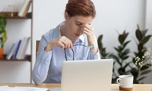 Kvinna sitter framför en vit laptop med en kopp kaffe vid sidan. Hon ser trött ut och tar sig för ögonen. 