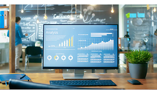 Datorskärm på ett kontor visar analyssida. Möte pågår i bakgrunden med stor blackboard.