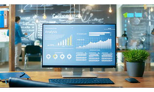 Datorskärm på ett kontor visar analyssida. Möte pågår i bakgrunden med stor blackboard. 