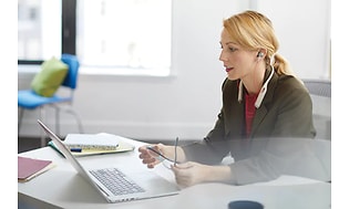 Kvinna i mörka kläder sitter med sin laptop vid ett vitt skrivbord och arbetar, hon har på sig in-ear hörlurar.