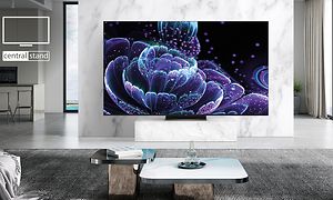 Stor TV-skärm med lila blomillustration som står på ett fotstativ i ett stort, ljust och öppet vardagsrum med stora fönster