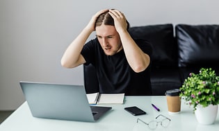 En frustrerad man som tar sig till huvudet medan han tittar på sin bärbara dator framför sig på skrivbordet