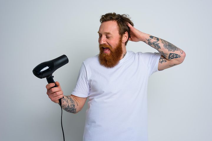 Glad man med vit t-shirt, skägg och tatueringar som sjunger in i en svart hårtork