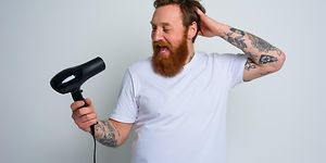 Glad man med vit t-shirt, skägg och tatueringar som sjunger in i en svart hårtork