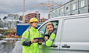 B2B - En kvinna med hjälm, ljusgul arbetsjacka och surfplatta pratar med en man i en bil på en byggarbetsplats