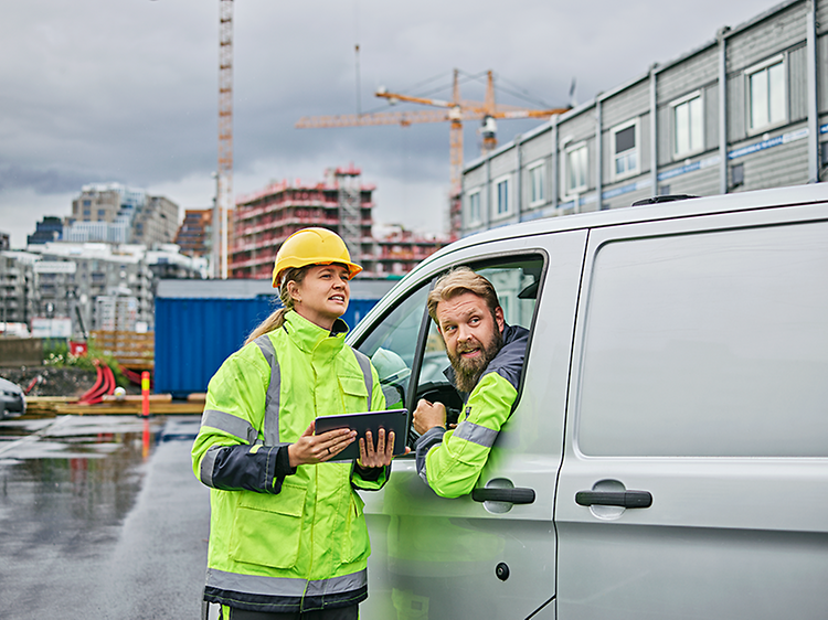 B2B - En kvinna med hjälm, ljusgul arbetsjacka och surfplatta pratar med en man i en bil på en byggarbetsplats