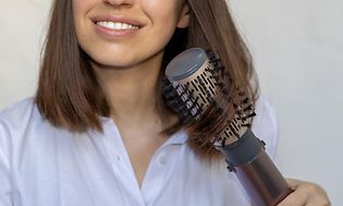 Ung kvinna som ler använder en varmluftsborste för att styla sitt bruna, halvlånga hår