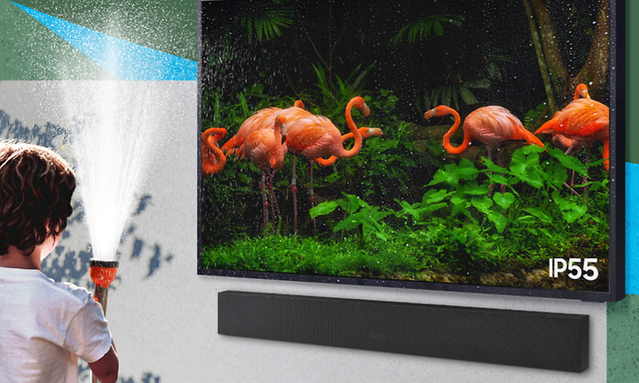 Pojke som står med en sprutande vattenslang framför en TV-skärm som visar flamingos på en vägg utomhus med en soundbar under