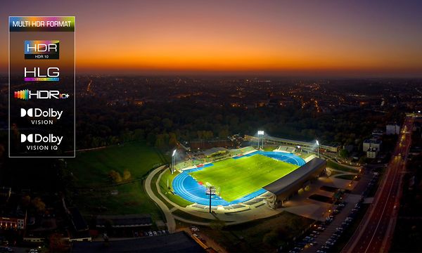 En upplyst fotbollsstadion i en storstad med mörk kvällshimmel med solnedgång i bakgrunden