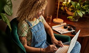 En glad kvinna med lockigt hår och tatueringar som sitter på ett café med sin HP 2-in-1 Chromebook i knät och hörlurar på bordet