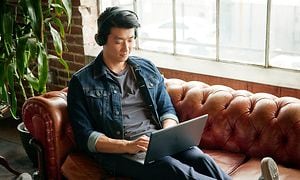En asiatisk man med hörlurar sitter i en soffa bredvid ett stort fönster med sin HP X360 Chromebook