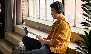 En man med asiatiskt utseende med gul jacka och hörlurar som sitter i en trappa vid ett fönster med sin HP X360 Chromebook