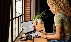 En kvinna med lockigt hår och rutig tröja arbetar vid sin HP 2-in-1 Chromebook bredvid ett stort fönster