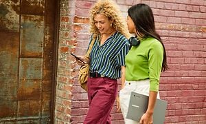 Två glada unga kvinnor går på en gata med en Samsung Galaxy Chromebook och en telefon i händerna