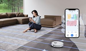 Kvinna som sitter på golvet och kontrollerar robotdammsugaren bredvid sig via sin smartphone och en bild av appen bredvid