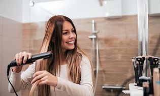 Glad kvinna med långt brunt hår som står i ett badrum vid en spegel och plattar håret