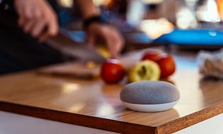 Google Nest mini på en köksbänk där någon skär upp äpplen i bakgrunden