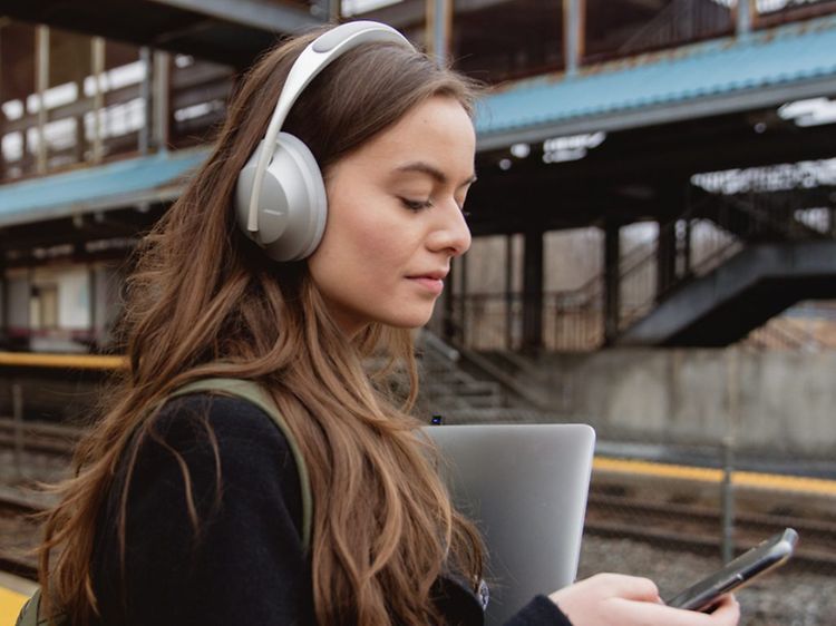 Kvinna med långt hår och over-ear-hörlurar på en folktom tågperrong tittar på sin mobiltelefon