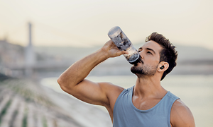 Vältränad man med grått linne och in-ears-hörlurar som står utomhus dricker ur en vattenflaska
