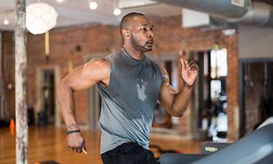 Svettig, färgad man med grått linne och in-ears i öronen springer på ett löpband på ett gym.