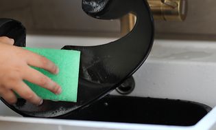 Person som rengör en airfryer med en grön disksvamp i en vit fyrkantig diskho med mässingkran i bakgrunden