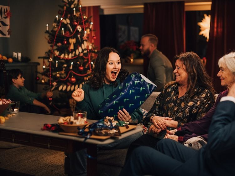 Familj som firar jul tillsammans i ett julpyntat vardagsrum och en ung kvinna som blir glad för en julklapp från Elgiganten
