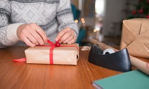 Kvinna med ljusgrå stickad tröja slår in julklappar med tejprulle och inslagningspapper bredvid sig på ett bord  