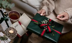 En kvinna med vitt nagellack och julklappar och en kaffekopp på ett bord bredvid sig håller en julklapp och ett kort i handen 