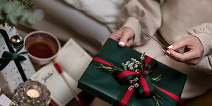 En kvinna med vitt nagellack och julklappar och en kaffekopp på ett bord bredvid sig håller en julklapp och ett kort i handen 