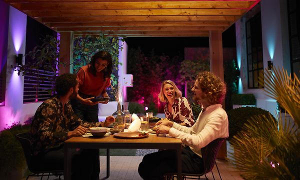 Två kvinnor och två män som äter vid ett bord utomhus på en altan på kvällstid mellan två hus som är upplysta med vägglampor