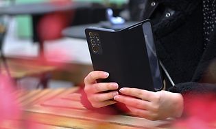 En kvinna håller en mobiltelefon med svart fodral med händerna på ett bord som står på en uteservering eller liknande