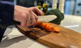 Kvinna skär upp morot på en skärbräda med broccoli i bakgrunden