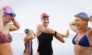 Kvinnliga simmare med simmössor i en grupp som står i vattnet utomhus där en av dem har fitbit-charge-4