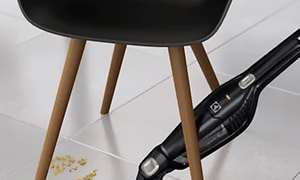En svart handhållen dammsugare suger upp matrester på golvet under en stol. .
