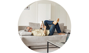 An older man lies on a grey couch reading a book