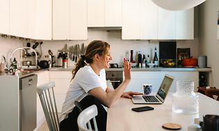 Woman sitting in front of a laptop waving to the webcam