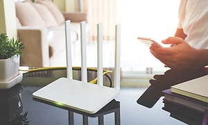 Vit router placerad på ett blankt bord. Man sitter och tittar på en mobiltelefon, en ljusrosa soffa syns i bakgrunden. 