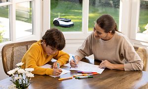 A mother and son drawing in the kitchen with Ecovacs GOAT robotic lawn mower in the background