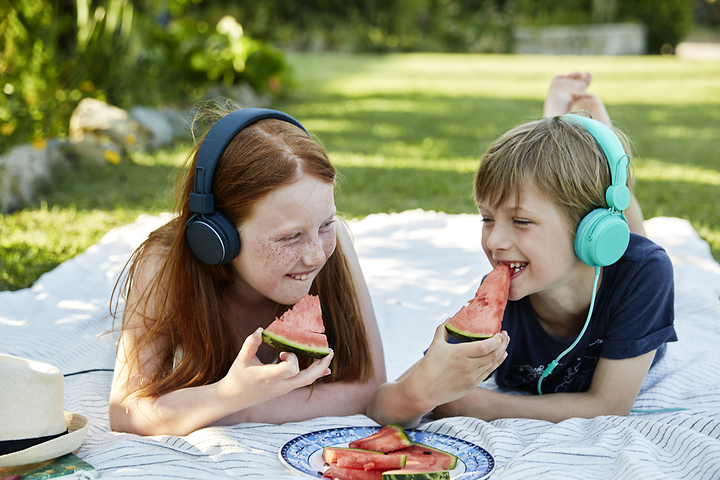 Two children lying on a blanket outside while eating watermelon and listening to something on their headphones