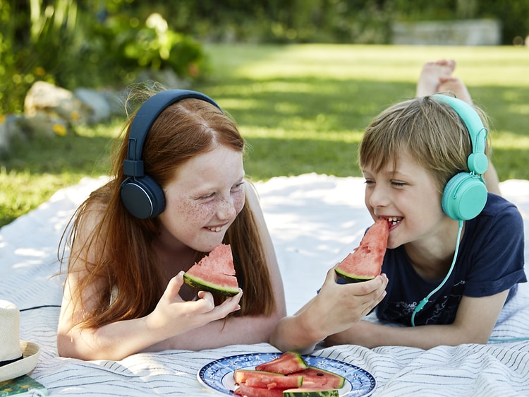 Two children lying on a blanket outside while eating watermelon and listening to something on their headphones