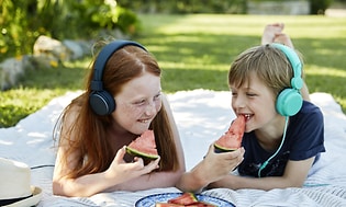 Two children lying on a blanket outside while eating watermelon and listening to something on their headphones